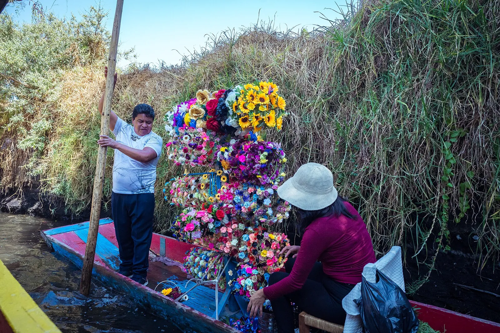 En sælgerbåd på en kanal i Xochimilco fyldt med bundter af kunstige blomster og blomsterkranse, med en bådfører stående agter