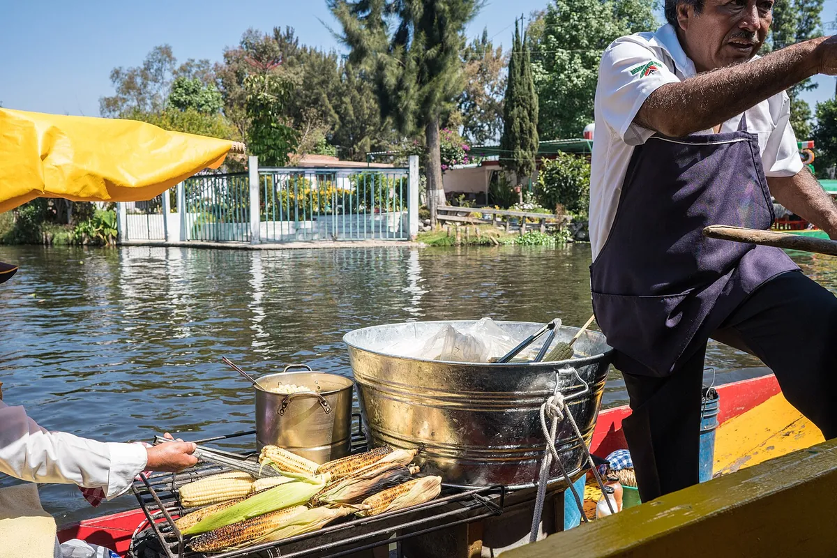En bådfører, der griller majs på en kulgrill på en lille båd i en kanal i Xochimilco, mens en kunde på en anden båd rækker over for at betale