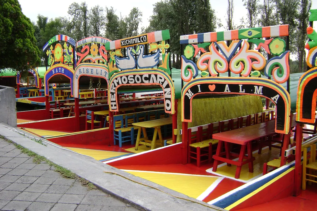 Malede trajineras med buede navneplader og træborde fortøjet ved Cuemanco-landingspladsen i Xochimilco