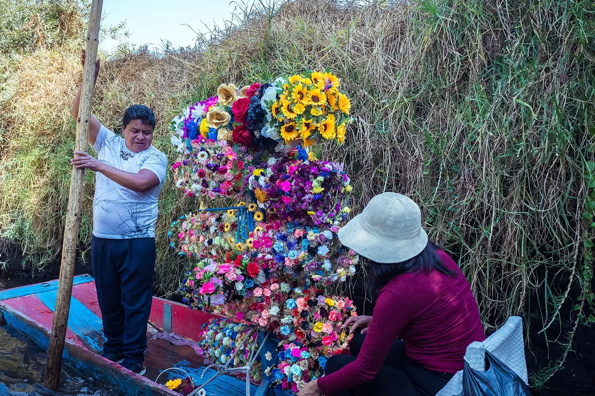 En sælger, der står agter i en båd fyldt med bundter af kunstige blomster og blomsterkranse i en kanal i Xochimilco