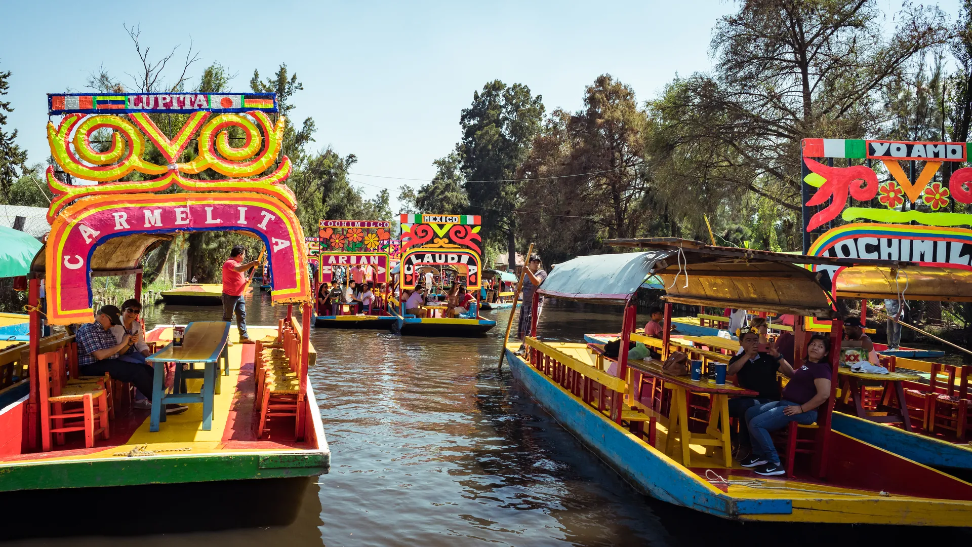 Brightly painted trajineras on a Xochimilco canal on a sunny day, with people seated at the long tables on board and more boats moored behind